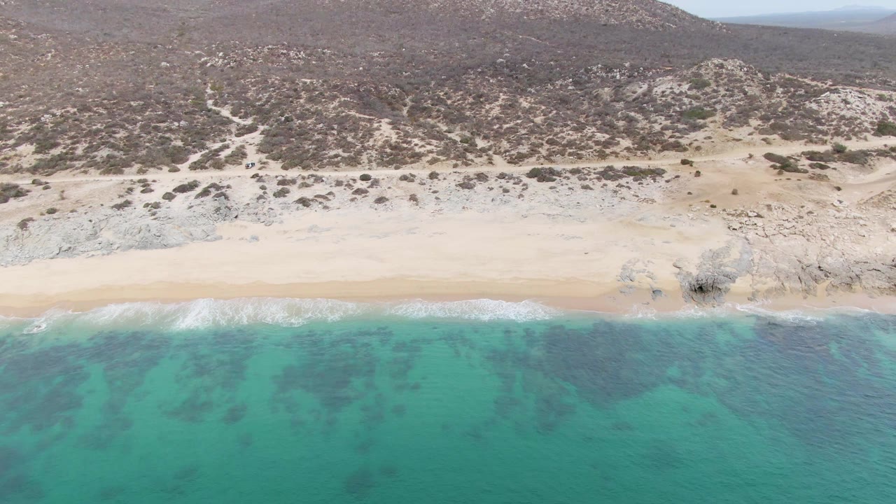 Aerial frame at Punta Gorda Beach, Baja California Sur, showing turquoise water in the foreground, a pale sand strip, rocky hillside terrain behind it, and no visible people.