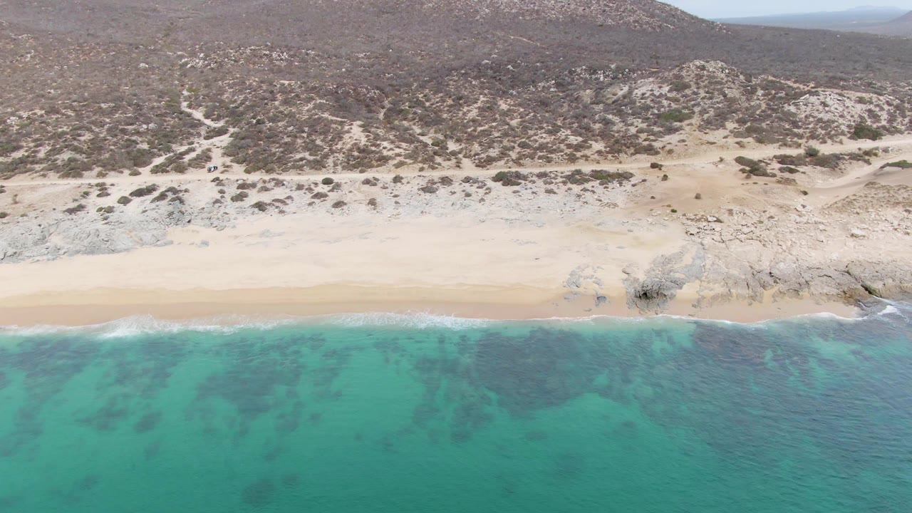 Aerial frame at Punta Gorda Beach where darker patches in the turquoise water sit below a clean sand band, with rocky desert hills across the background and no visible people.