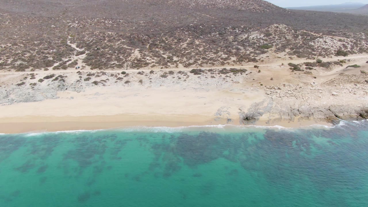 Aerial shoreline view with turquoise water, foam, sand, and dry hills.