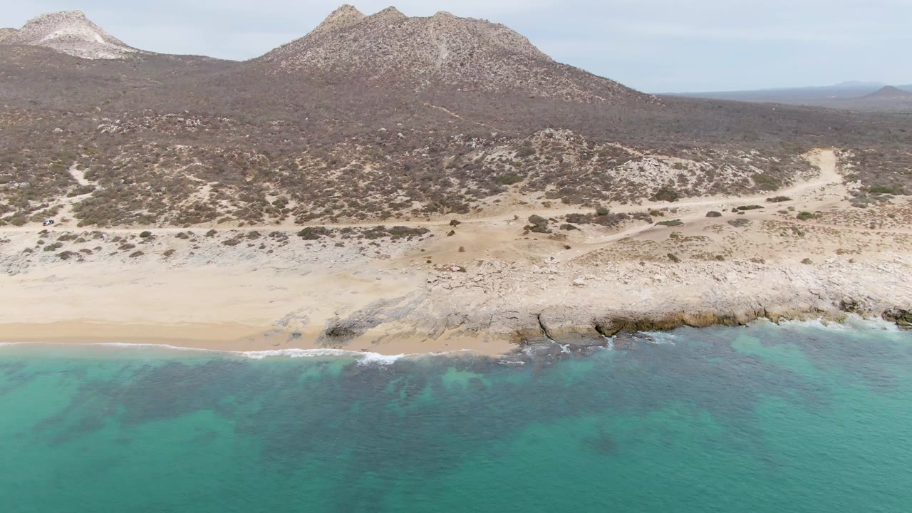 Aerial frame over Punta Gorda Beach in Baja California Sur, with turquoise water in the foreground, a pale sandy beach and rocky shelf at shore, scrubby desert hills behind, and no visible people.