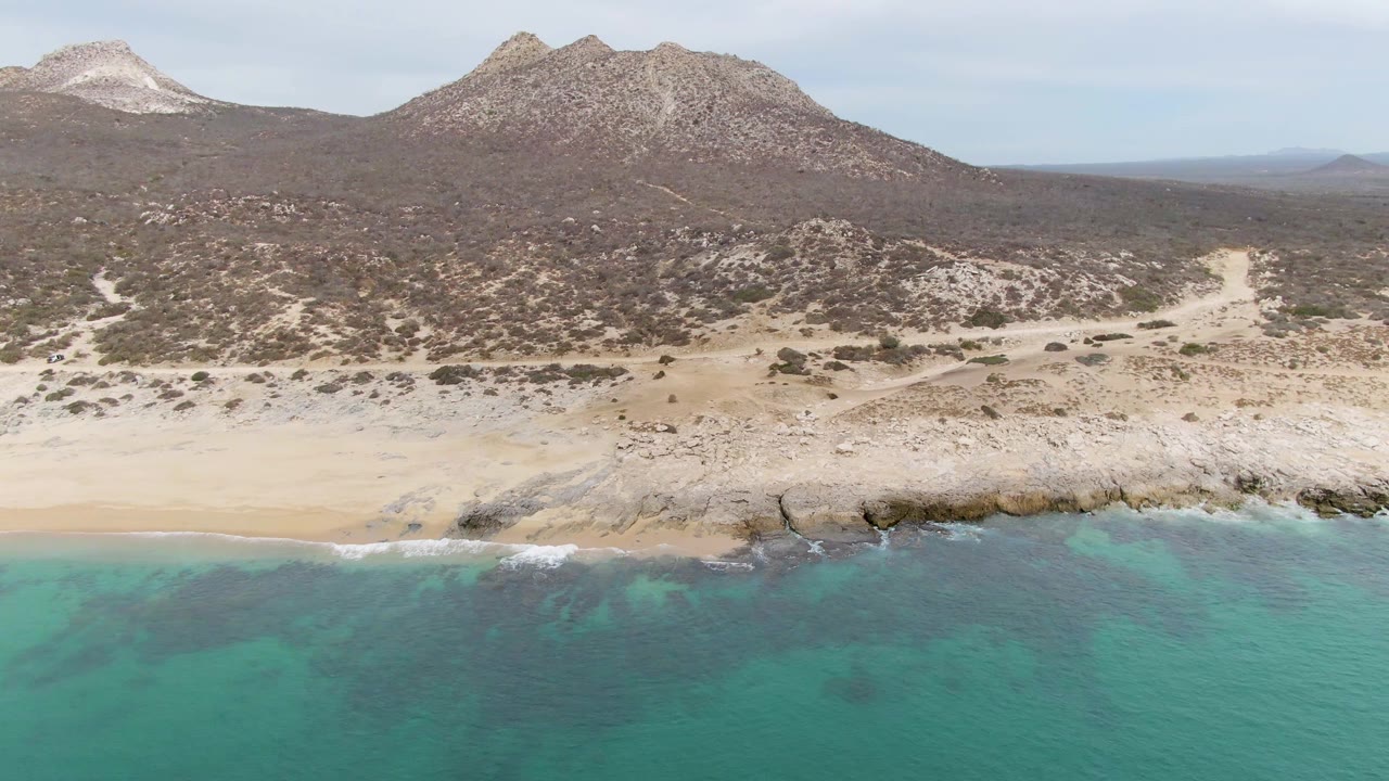 Wide drone view of a pale beach below rocky desert hills and mountains.