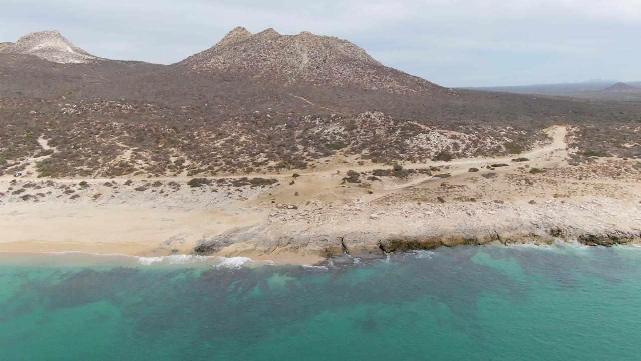 The aerial angle over Punta Gorda Beach shows turquoise water reaching a jagged rocky shoreline, with sparse desert scrub and two rugged mountain forms beyond it and no visible people.