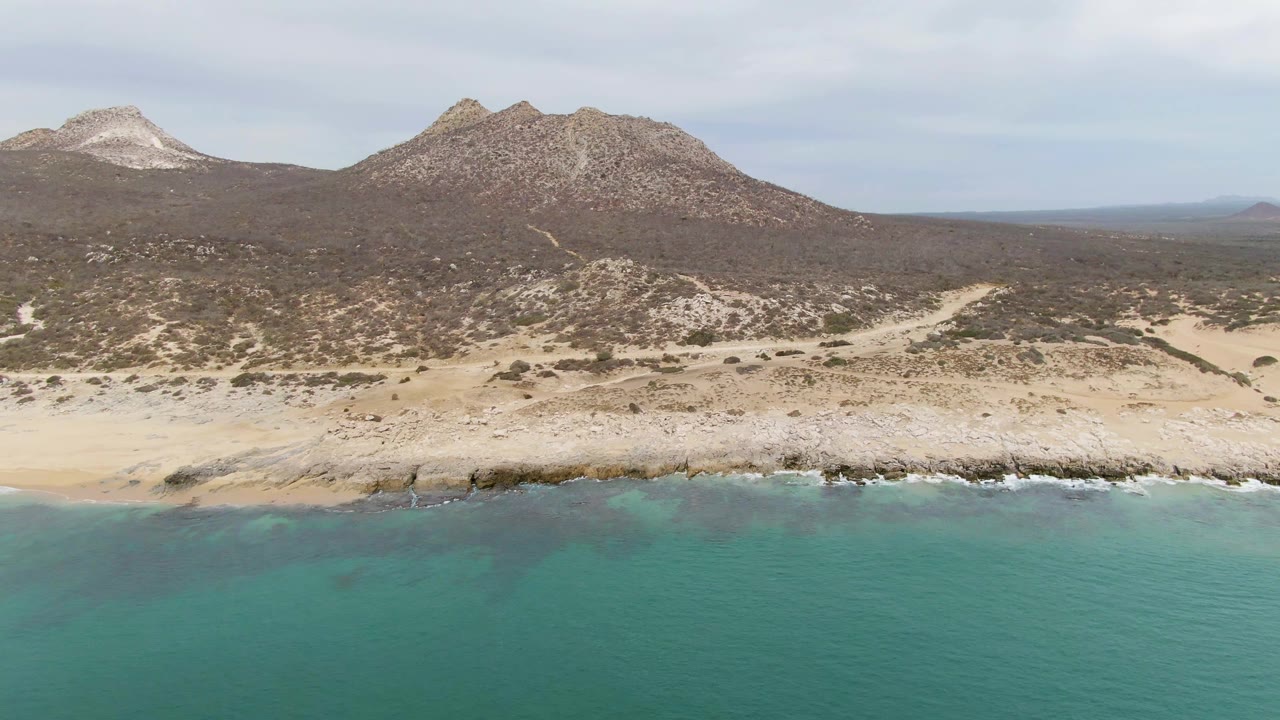Aerial view of turquoise ocean, sandy rocky shore, and coastal hills.