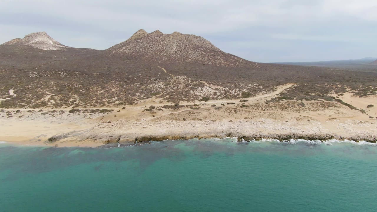 Aerial frame at Punta Gorda Beach showing slightly shifted wave texture along the rocky shoreline, dry hills in the background, pale cloud cover, and no visible people.
