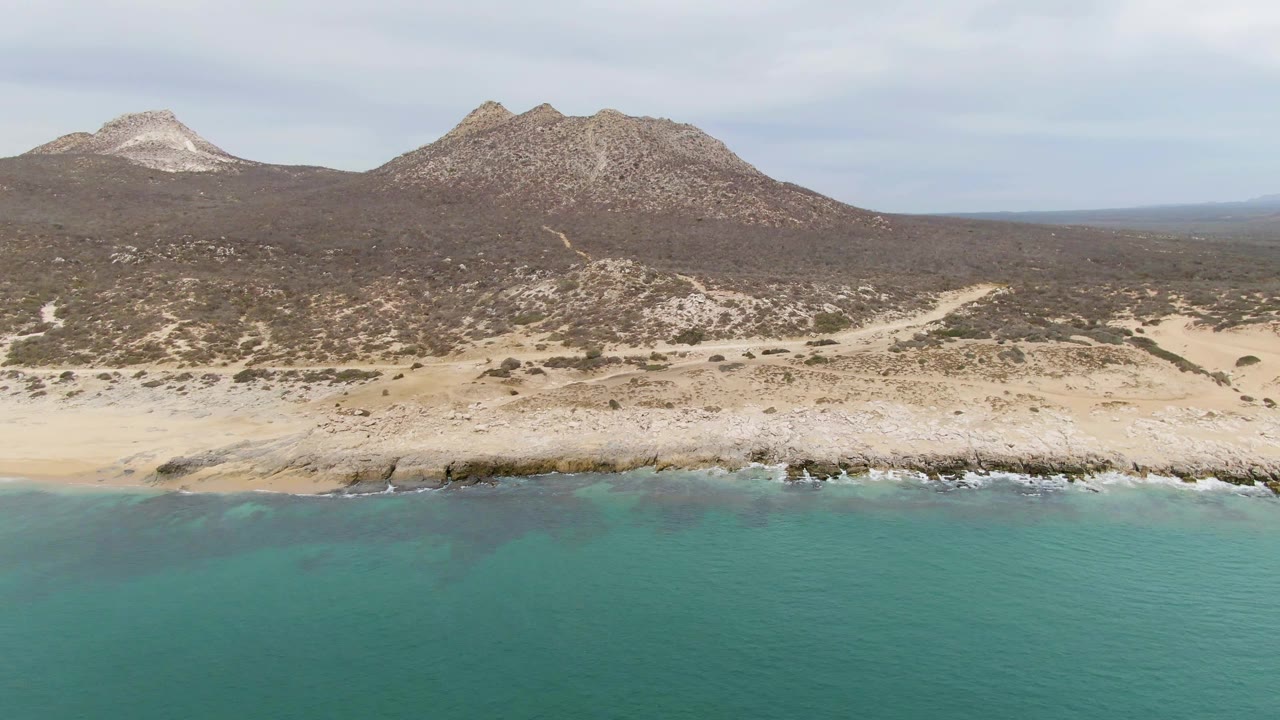 Mid-clip aerial view of Punta Gorda Beach with calm teal ocean across the lower frame, a thin sandy and rocky shore, rugged hills behind it, and no visible people.