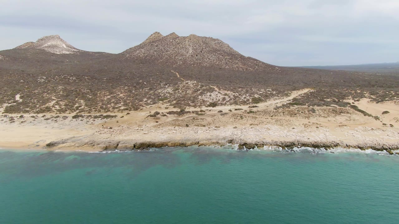 Aerial frame over Punta Gorda Beach with the water band lower in frame, a rocky pale shoreline across the center, scrubland leading to hills, and no visible people.
