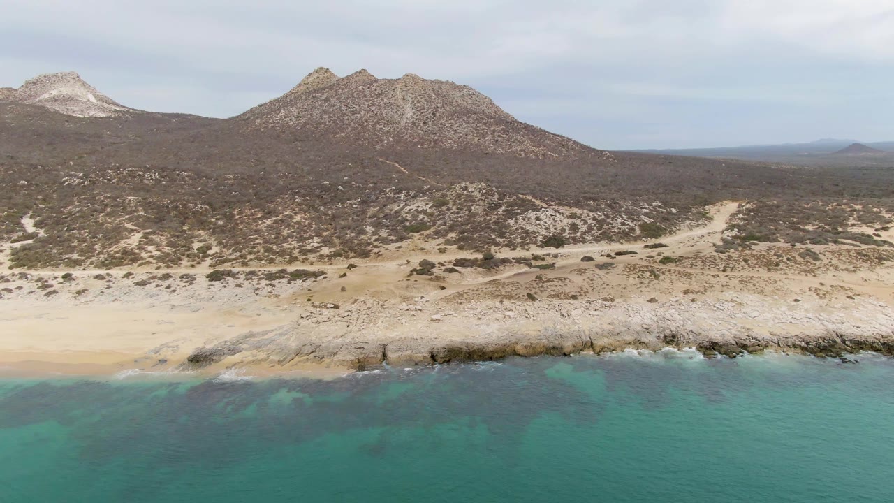 At Punta Gorda Beach in Baja California Sur, turquoise water meets a pale sandy and rocky shoreline below scrub-covered hills, with no visible people.