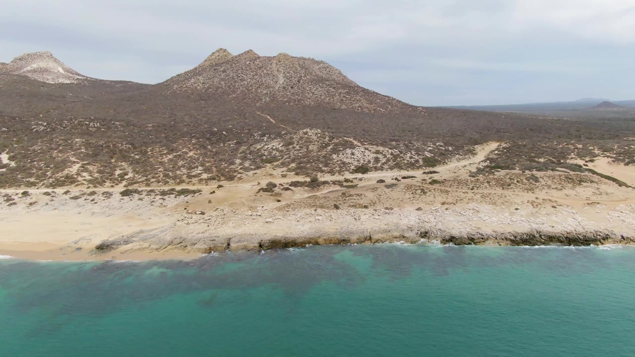 Aerial coastal view with waves, sand, desert scrub, and hills.