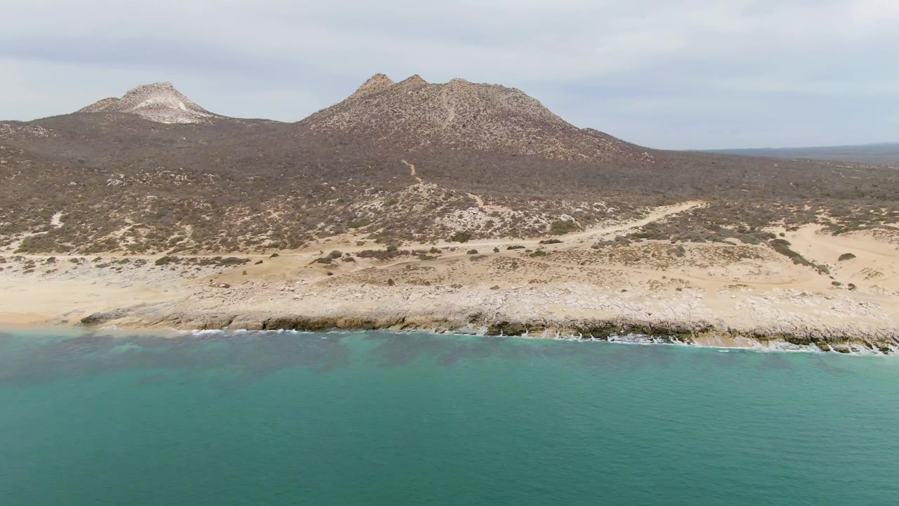 Turquoise ocean beside rocky sand shore with arid mountains behind.