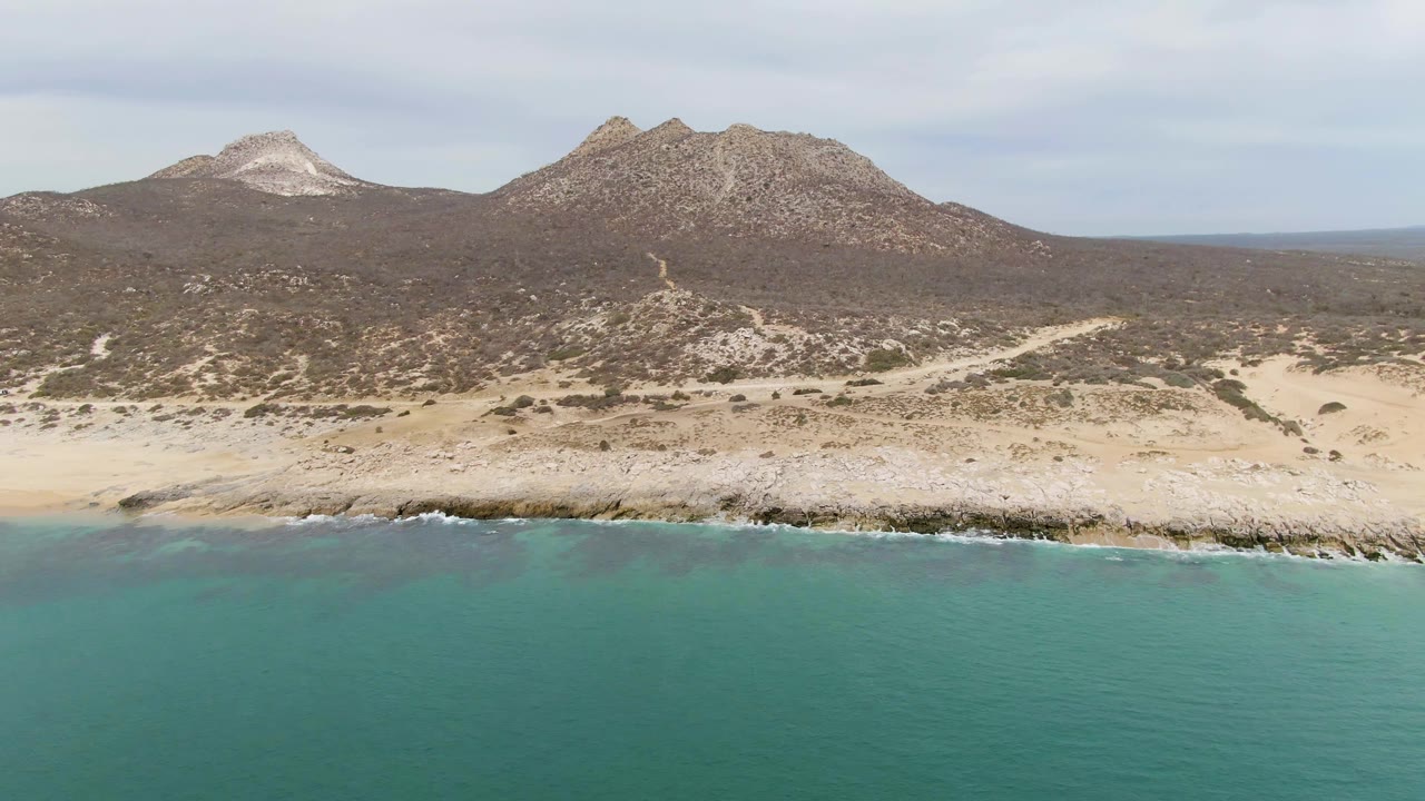 Aerial view at 0:01 over Punta Gorda Beach BCS, Mexico, showing calm turquoise water, thin surf on the rocky beach, tan desert ground, mountain ridges, and no visible people.