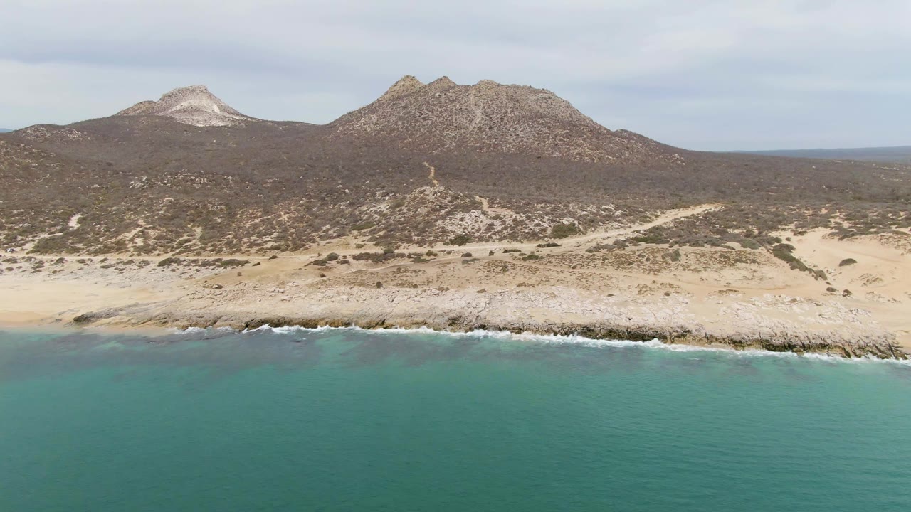 Mid-clip aerial frame of Punta Gorda Beach BCS, Mexico, with the ocean foreground, pale beach and rocks across the center, two mountain forms inland, muted cloud cover, and no visible people.