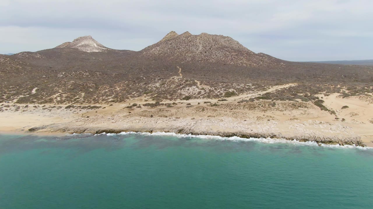 At 0:03, Punta Gorda Beach BCS, Mexico, appears from above with turquoise sea below, a broken white surf line, rocky sand, dark scrubland, rounded mountains, and no visible people.