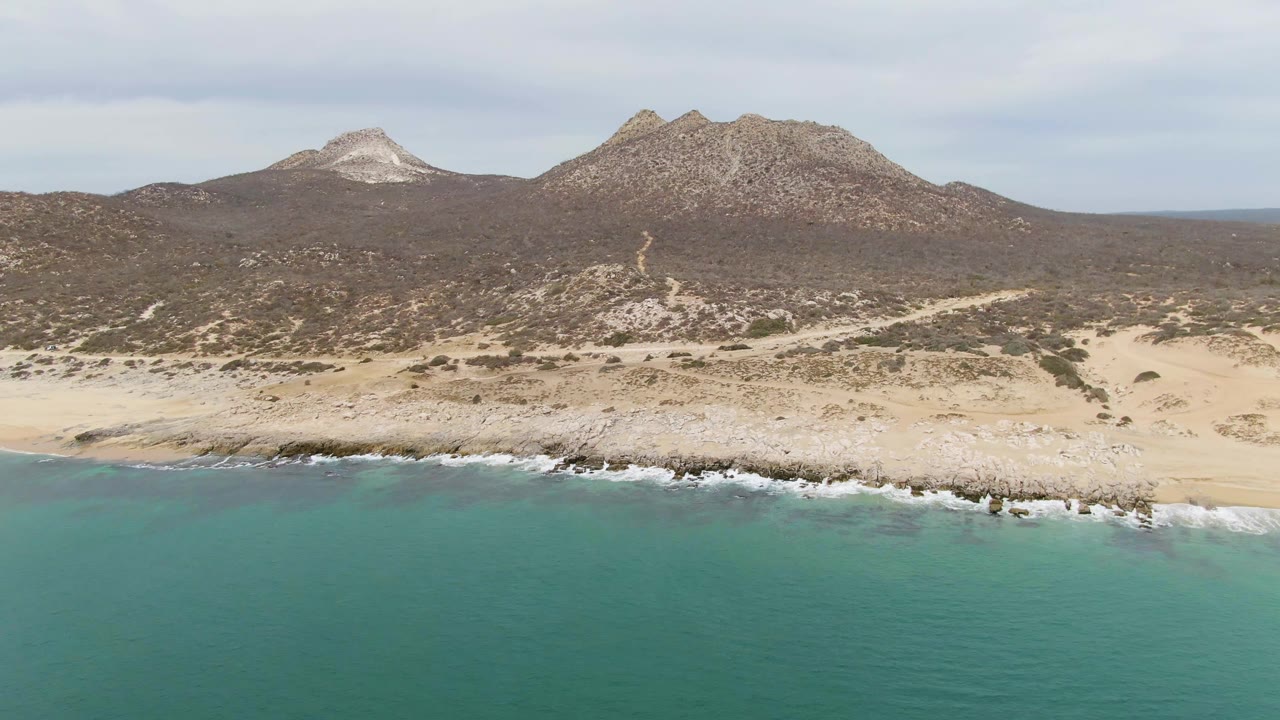 Opening aerial frame over Punta Gorda Beach in Baja California Sur, with turquoise ocean in the foreground, white surf on a sandy rocky shore, dry scrubland, low mountains, and no visible people.