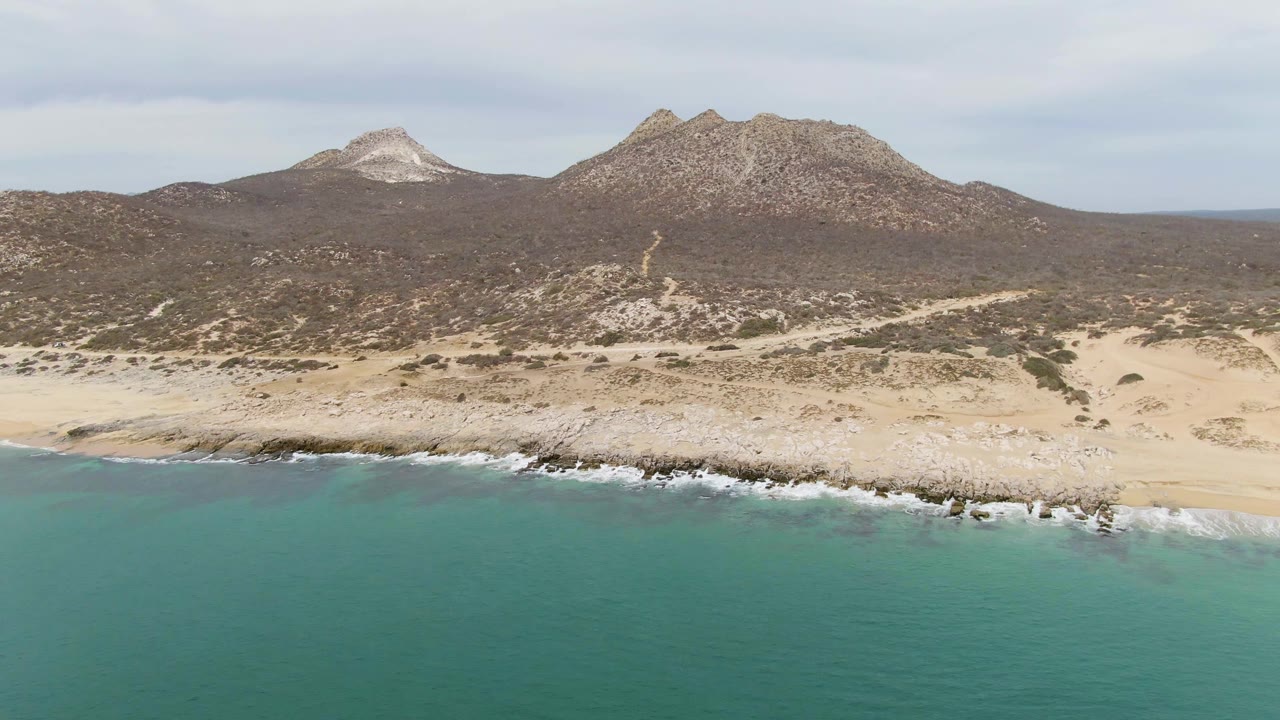 Aerial view at 0:01 over Punta Gorda Beach, showing blue-green water, a broken white wave line on the rocky sand, pale dunes, sparse vegetation, mountain ridges, and no visible people.