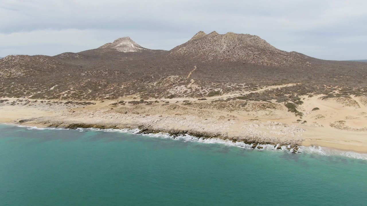 Aerial beach scene with teal water, foamy waves, dry dunes, scrub, and mountains.