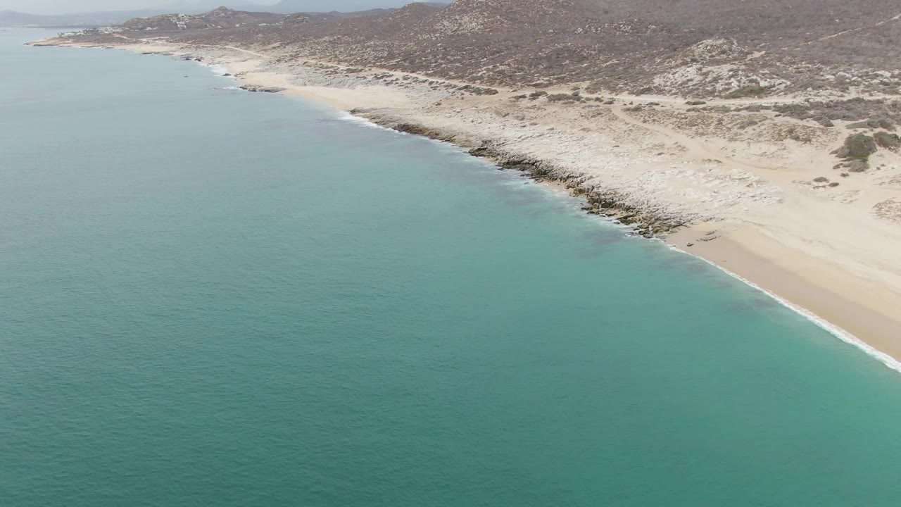 Aerial frame over Punta Gorda Beach in Baja California Sur showing turquoise water, pale sand, rocky shoreline, dune scrub, distant hills, and no visible people.