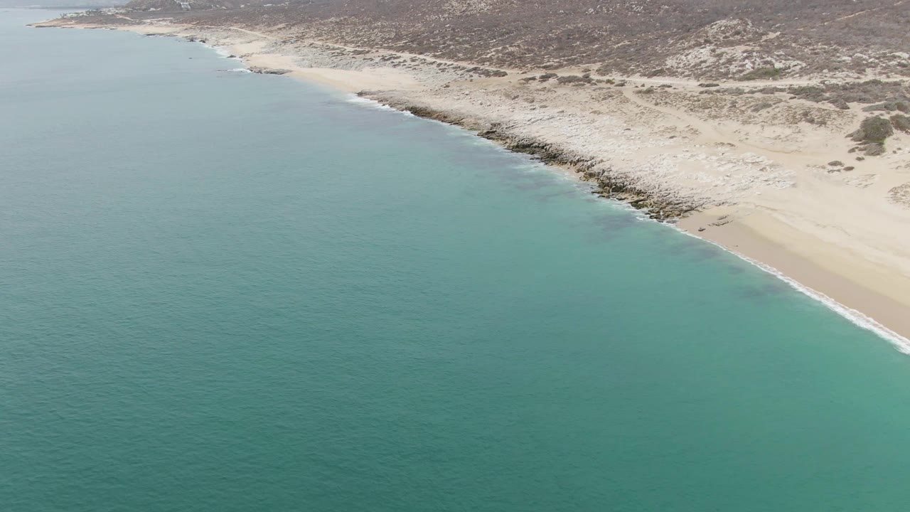 Aerial frame over Punta Gorda Beach with the turquoise sea extending across the foreground, a narrow surf line, pale beach, rocky edge, dunes, and no visible people.
