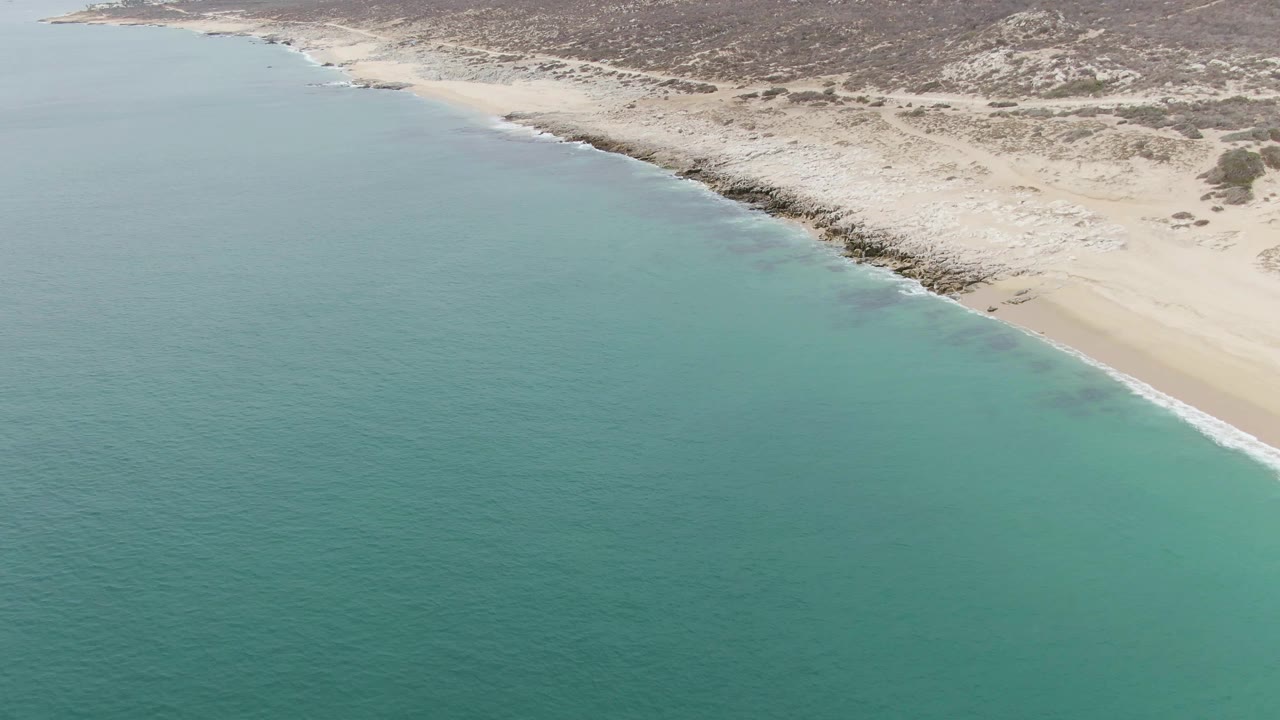 Aerial frame at Punta Gorda Beach showing the shoreline running diagonally across calm turquoise water, with sand, rocks, scrub dunes, hills, and no visible people.