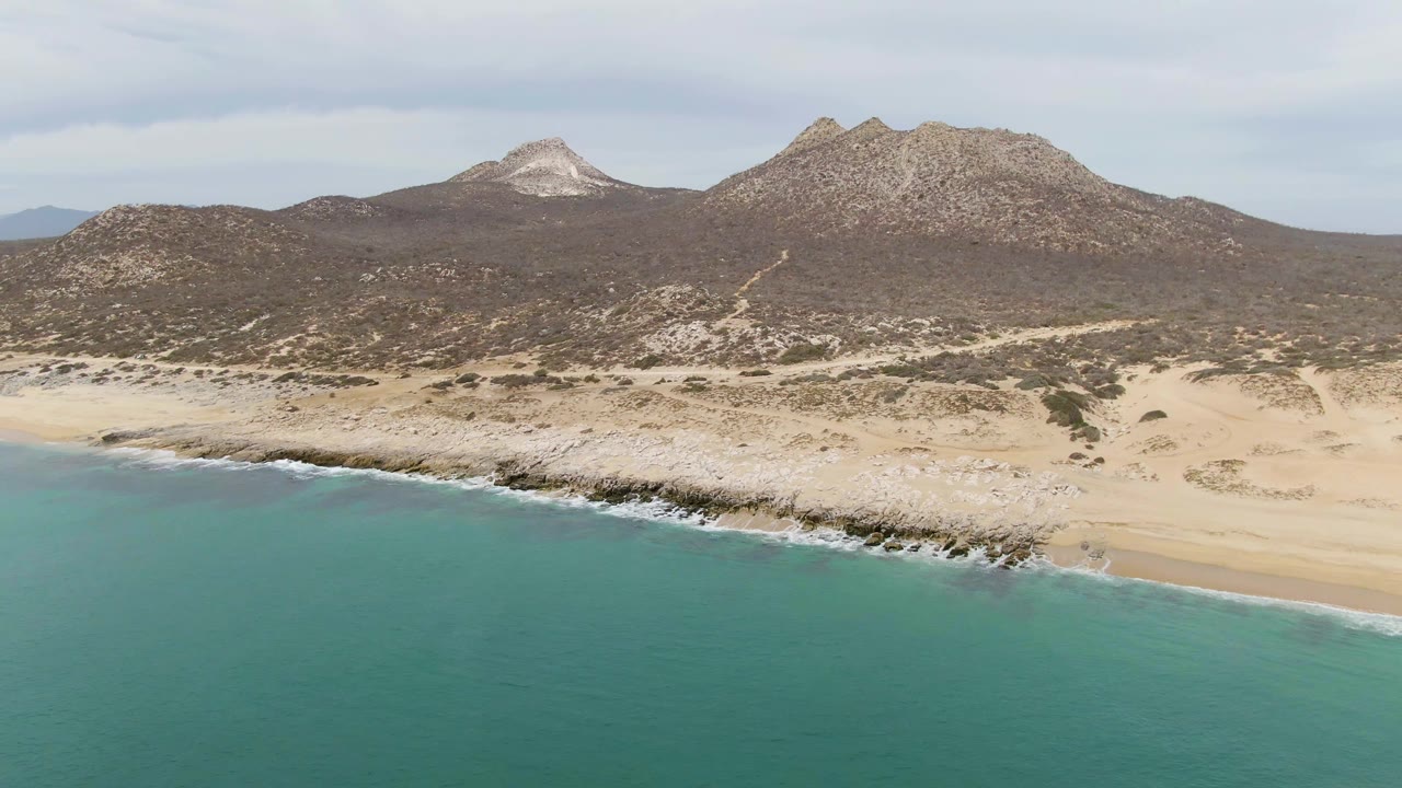 Aerial frame from Punta Gorda Beach, Baja California Sur, showing turquoise ocean in the foreground, a pale beach and rocky shelf, and arid mountains beneath a cloudy sky.