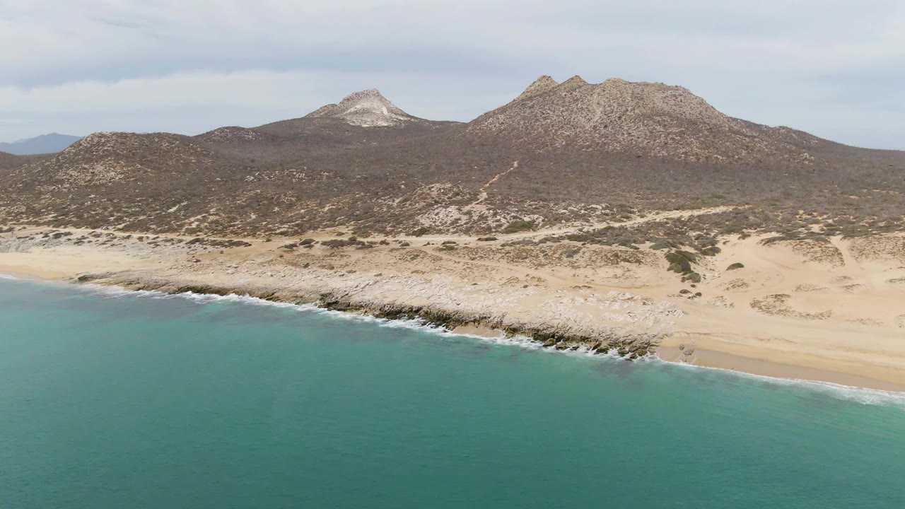 Wide aerial coastline with turquoise ocean, pale sand, rocky edge, and hills behind.