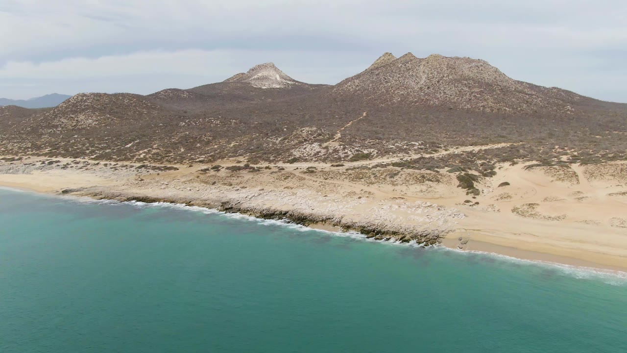Aerial view over Punta Gorda Beach with smooth turquoise water, a thin white wave line, sandy beach, rocky coastal band, and scrub-covered mountains in the distance.