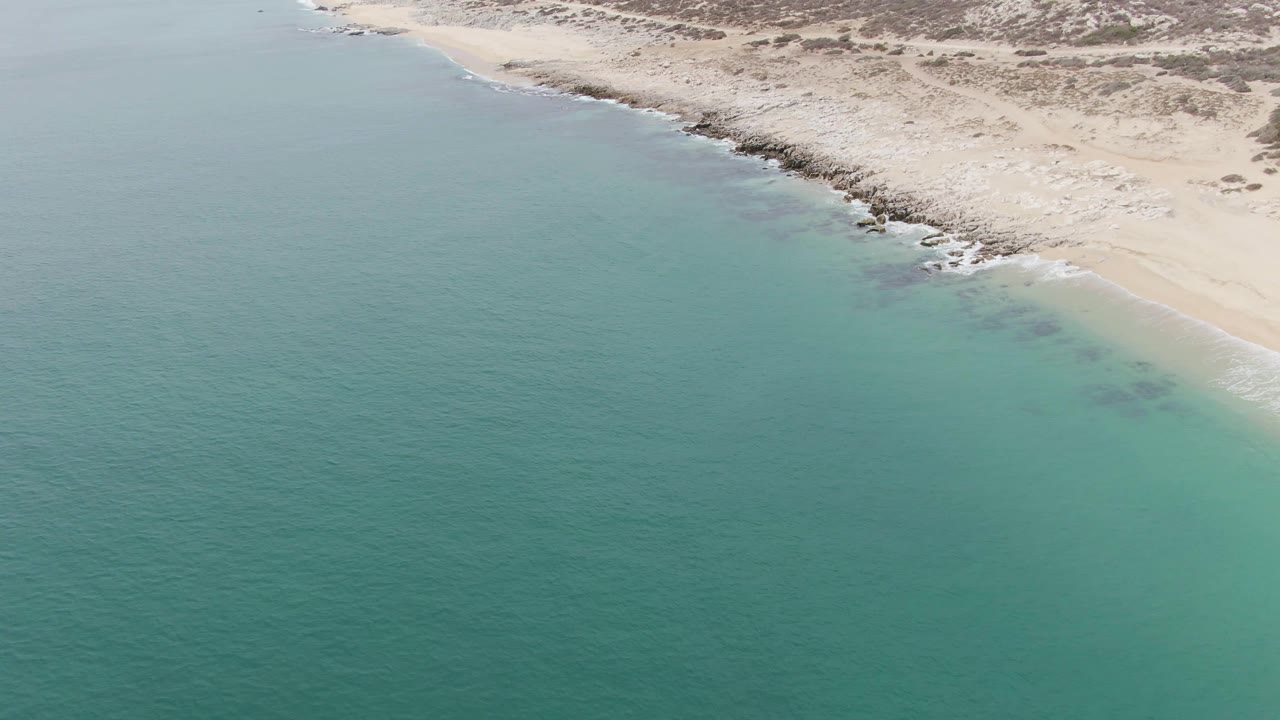 Aerial view over Punta Gorda Beach in Baja California Sur, with turquoise water in the foreground, pale sand and rocks along the upper shore, and no visible people.