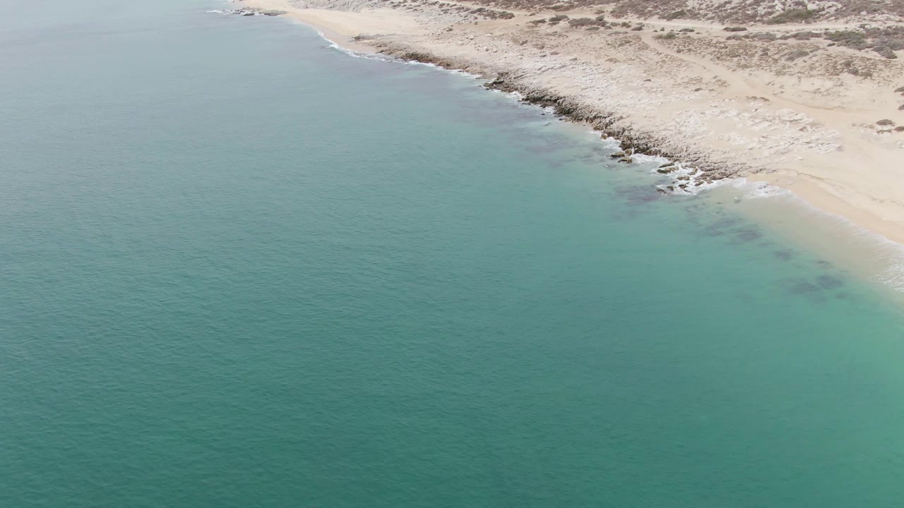 Aerial frame from Punta Gorda Beach showing a broad field of turquoise water, a narrow line of white surf, pale sand, dark rocks, and no visible people.
