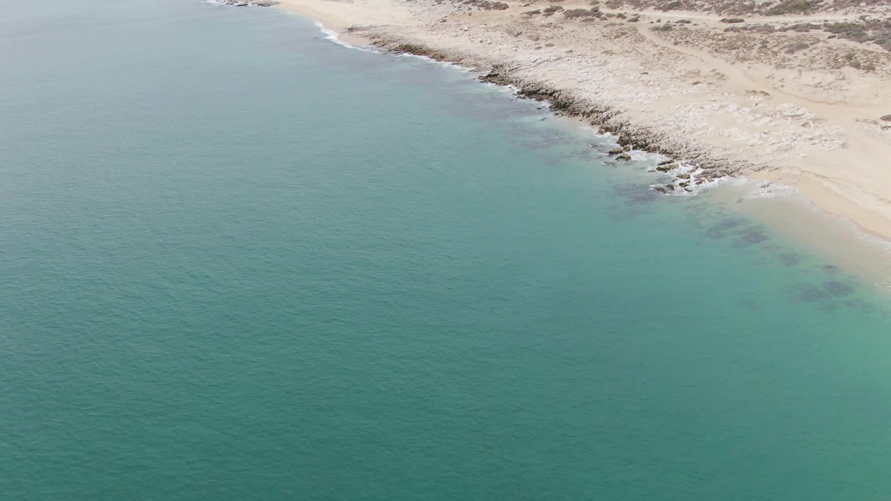 Aerial view at Punta Gorda Beach with blue-green water below, darker submerged patches near shore, sand and scattered rocks along the top edge, and no visible people.