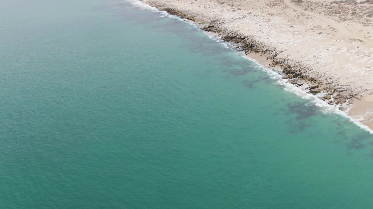 Aerial frame over Punta Gorda Beach with green-blue water, small white surf, and a rocky sandy beach edge continuing across the upper frame, no visible people.