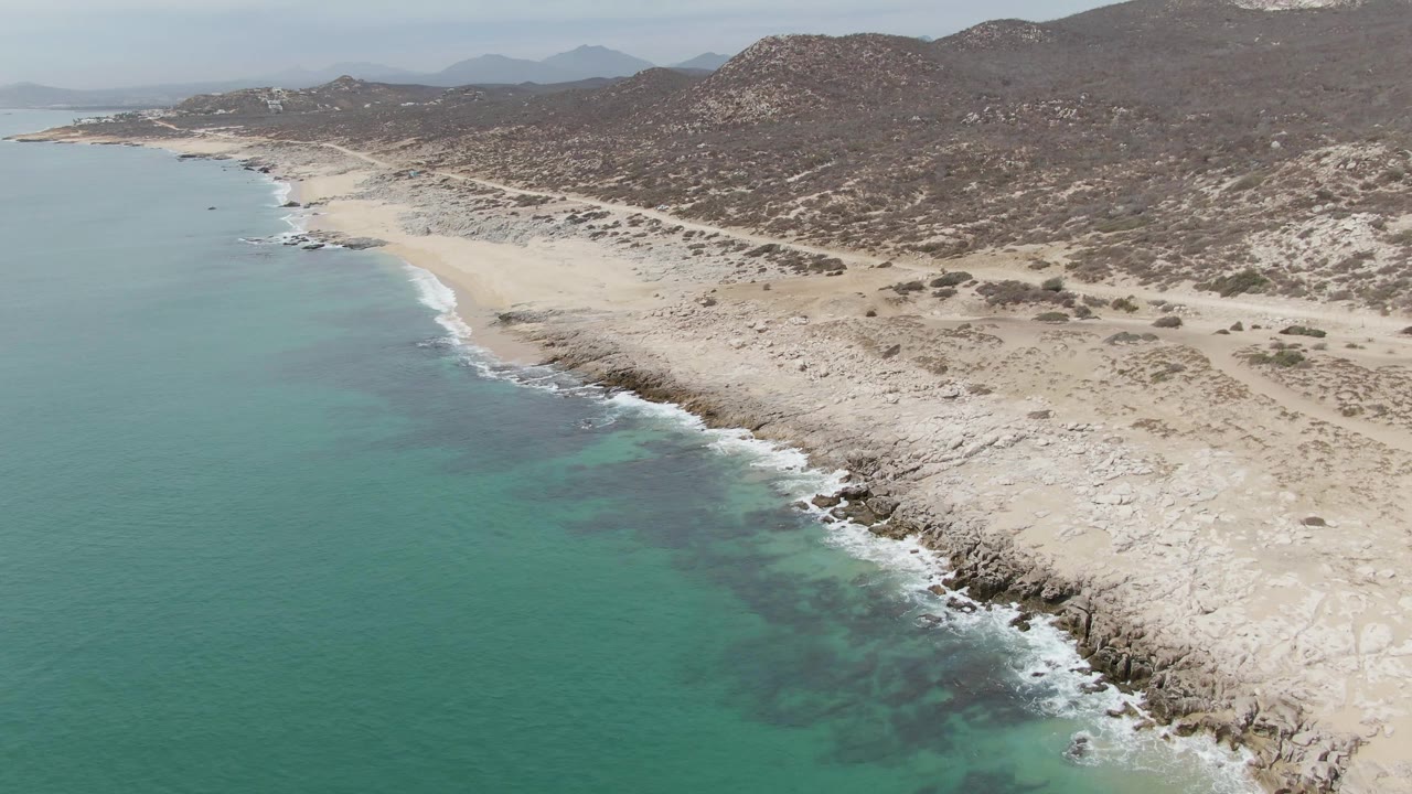 Aerial frame of Punta Gorda Beach showing more of the rocky shore and shallow turquoise water, with pale sand and scrub-covered hills inland and no visible people.