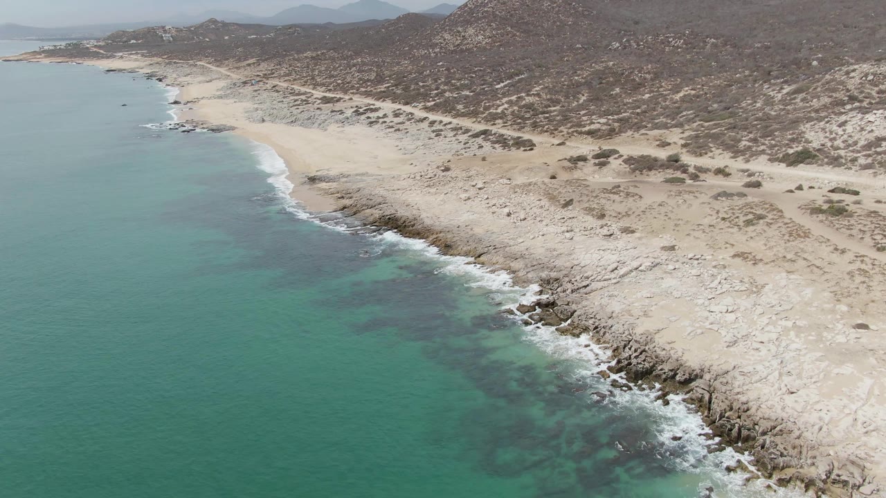 Aerial frame over Punta Gorda Beach with turquoise shallows filling the left side, white surf tracing the rocky shoreline, pale sand inland, and no visible people.