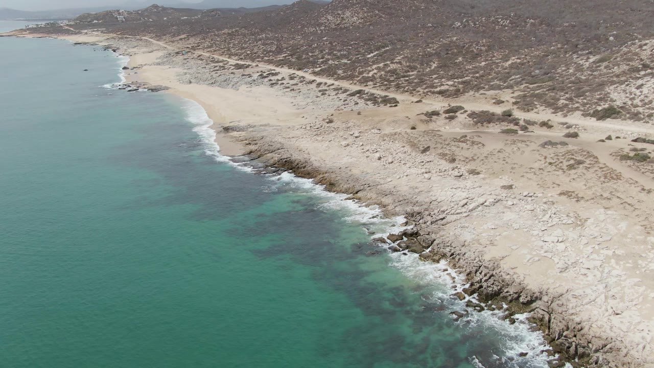 Aerial frame of Punta Gorda Beach from later in the clip, with open turquoise water along the left edge, a pale rocky beach crossing the frame, dry hills above, and no visible people.