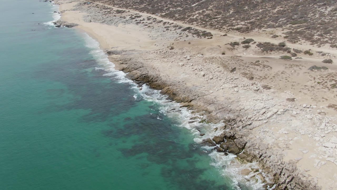 Aerial frame from Punta Gorda Beach, Baja California Sur, showing turquoise water on the left breaking into white foam along pale rock shelves and sand, with no visible people.