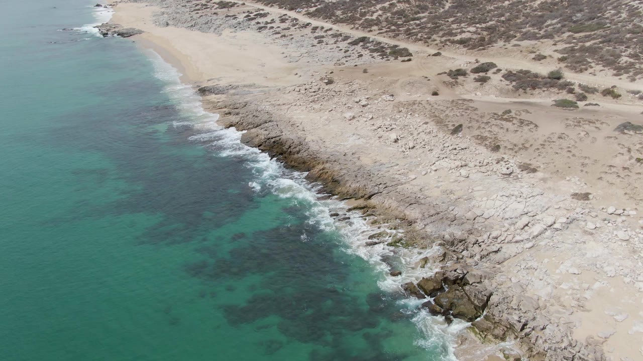 Aerial frame from Punta Gorda Beach, Baja California Sur, with dark nearshore patches under turquoise water, foam tracing the rocks, and pale beach and scrub above the coast, with no visible people.