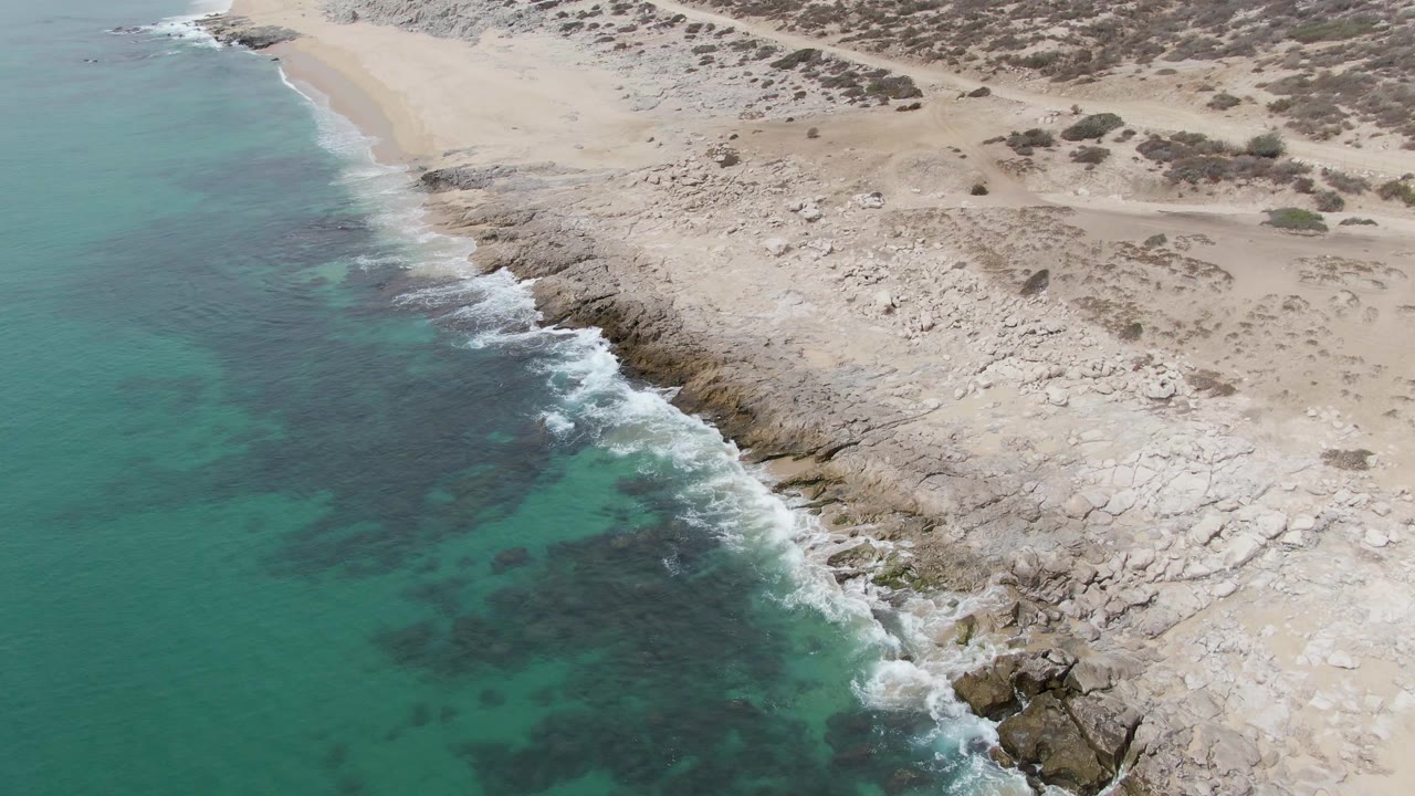 Aerial frame from Punta Gorda Beach, Baja California Sur, where the diagonal rock edge catches a line of white foam below sand and scattered low scrub, with no visible people.
