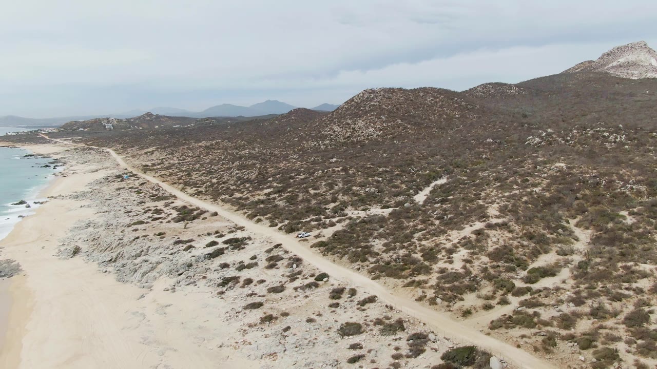 Aerial view of Punta Gorda Beach with a small white vehicle on the dirt road near the lower right, beach and ocean to the left, and scrubby hills beyond, with no visible people.