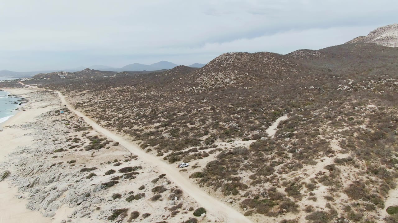 Aerial view of a dirt road beside sandy coast and scrub-covered hills.