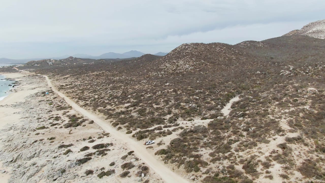 Aerial frame at Punta Gorda Beach, Baja California Sur with the vehicle lower on the dirt road, beach and water to the left, scrubland ahead, and no visible people.