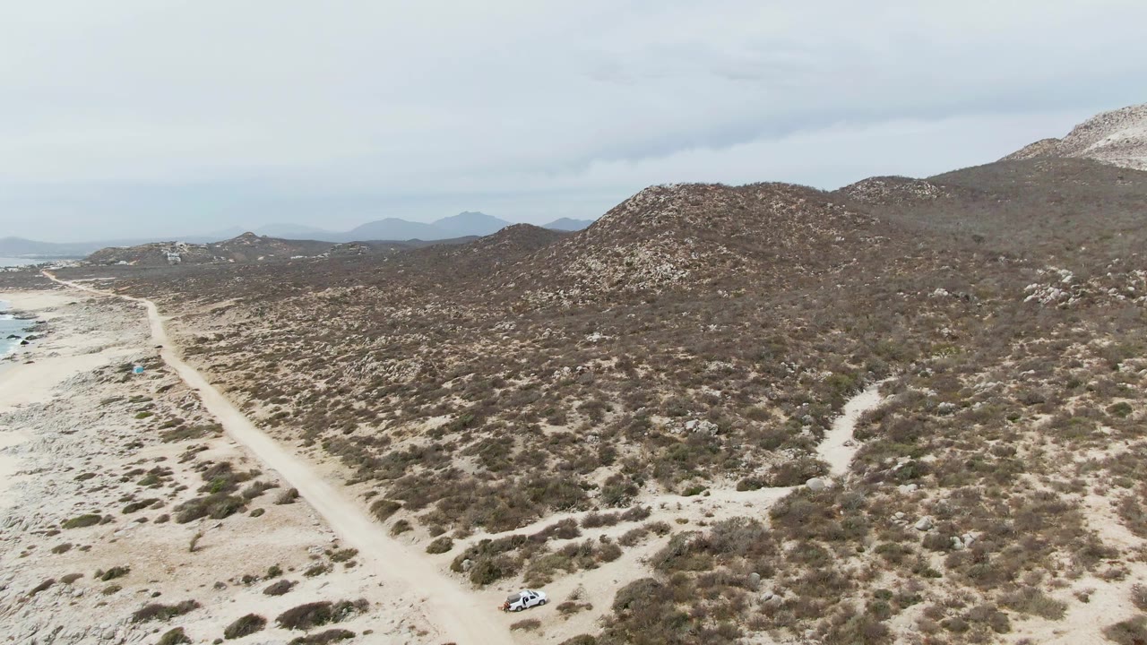 Aerial frame at Punta Gorda Beach, Baja California Sur with the sandy road nearer the left edge, scrub-covered slopes across the center, distant hills, and no visible people.