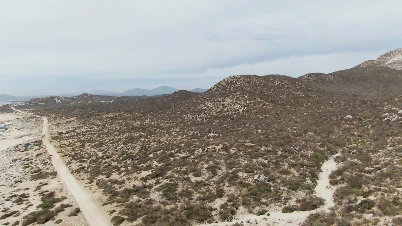 Aerial frame from Punta Gorda Beach, Baja California Sur, showing a pale dirt road along sandy scrub terrain with rocky hills ahead and no visible people.