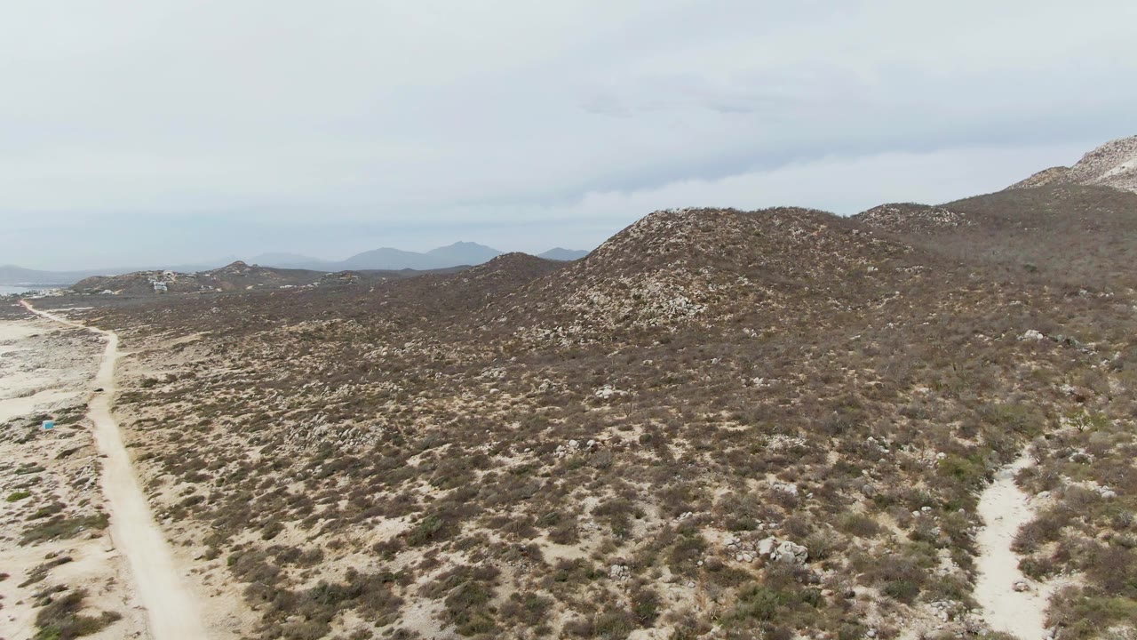 At 0:02, the aerial view over Punta Gorda Beach terrain shows the road bending through low desert vegetation, with brown hills and hazy distant ridges beyond and no visible people.