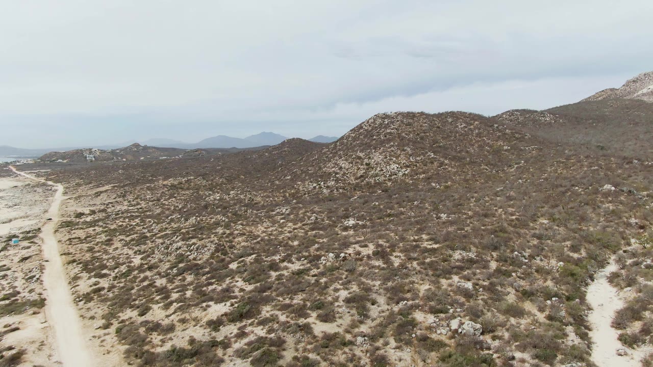 At 0:03, the Punta Gorda Beach clip shows the dirt track closer to the left edge, with scrubland stretching across the foreground and rocky hills under pale cloud cover, with no visible people.