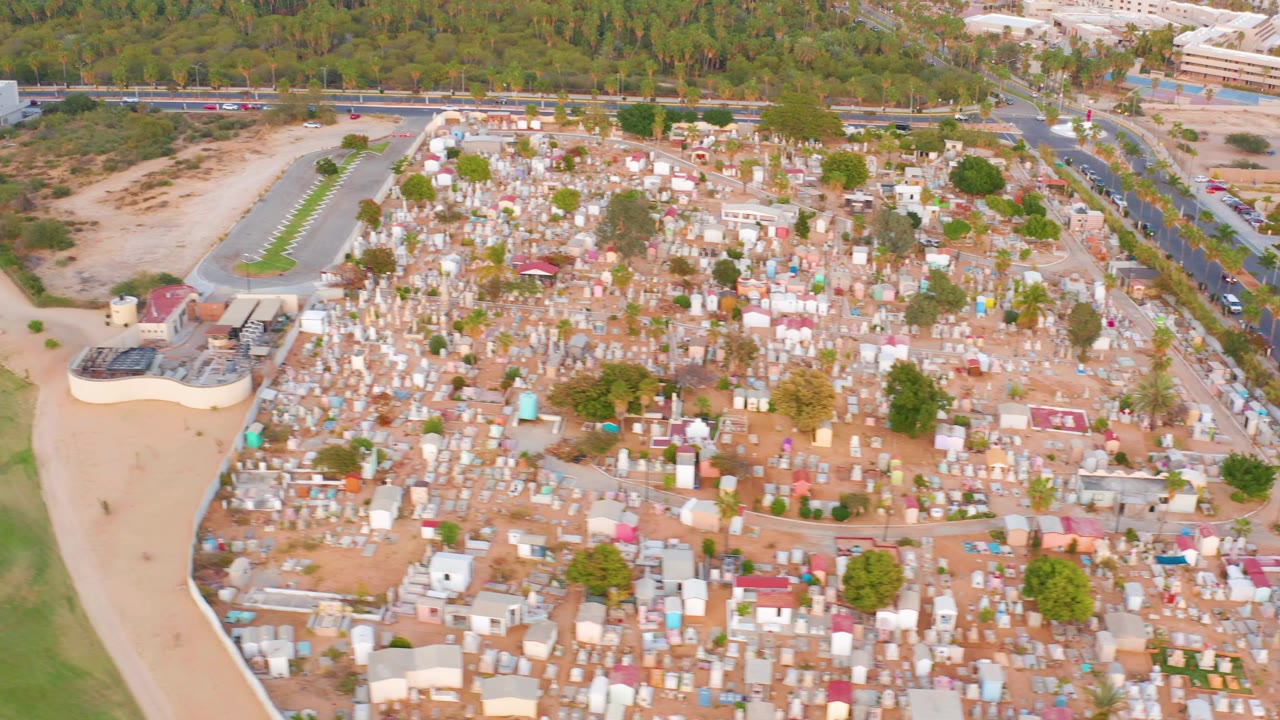 Opening aerial frame over dense small structures in San Jose del Cabo, Mexico, with a green field on the left, a curved dirt path, roadside vehicles in back, and no visible people.
