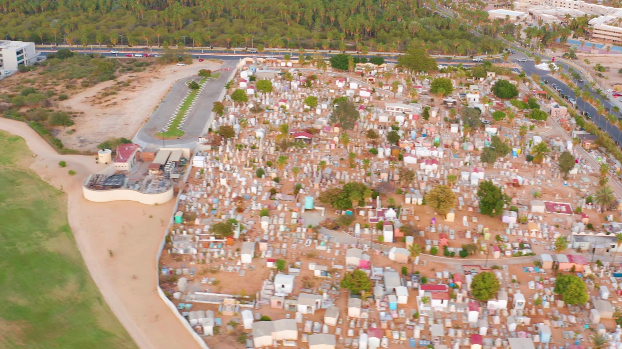 Aerial frame in San Jose del Cabo, Mexico showing the packed settlement centered in view, with open dirt lots, a road with vehicles, tree cover in the background, and no visible people.
