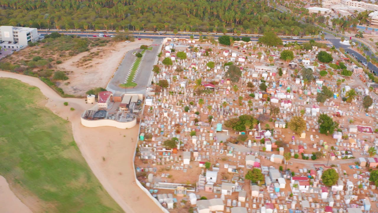 Aerial view in San Jose del Cabo, Mexico with dense light-colored structures on the right, green open land and a dirt path on the left, a paved road behind, and no visible people.