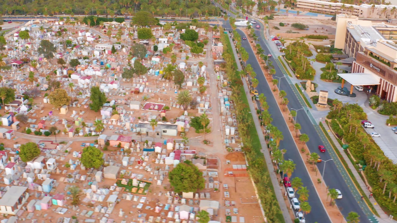 Overhead view of a large sandy cemetery in San Jose Del Cabo, Mexico, with many small graves, bordering roadways, a large building at right, and no visible people.