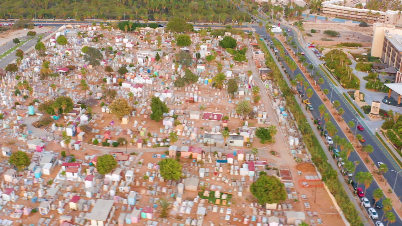 High aerial view of the cemetery in San Jose Del Cabo with dense graves across sandy ground, landscaped medians, cars on adjacent roads, and no visible people.