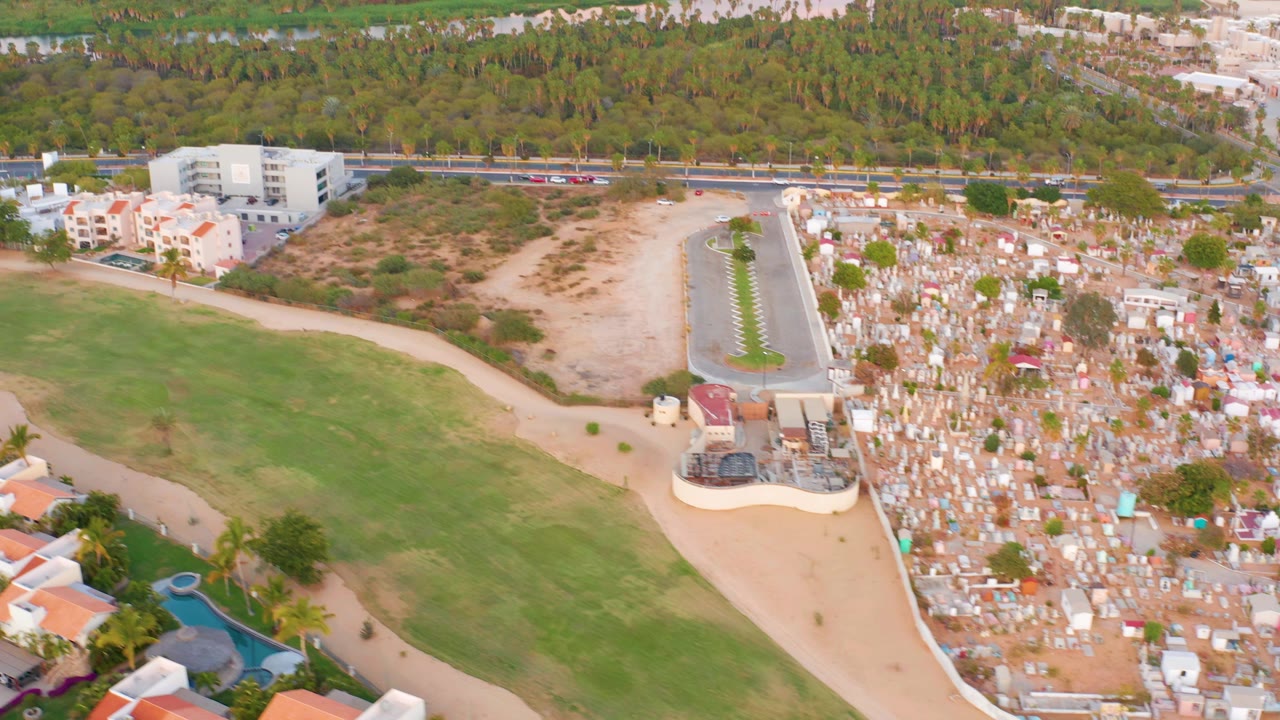 Aerial frame over San Jose del Cabo showing a green field, red-roofed residential buildings, a cemetery area, a roadside with cars, beach, water, and no visible people.