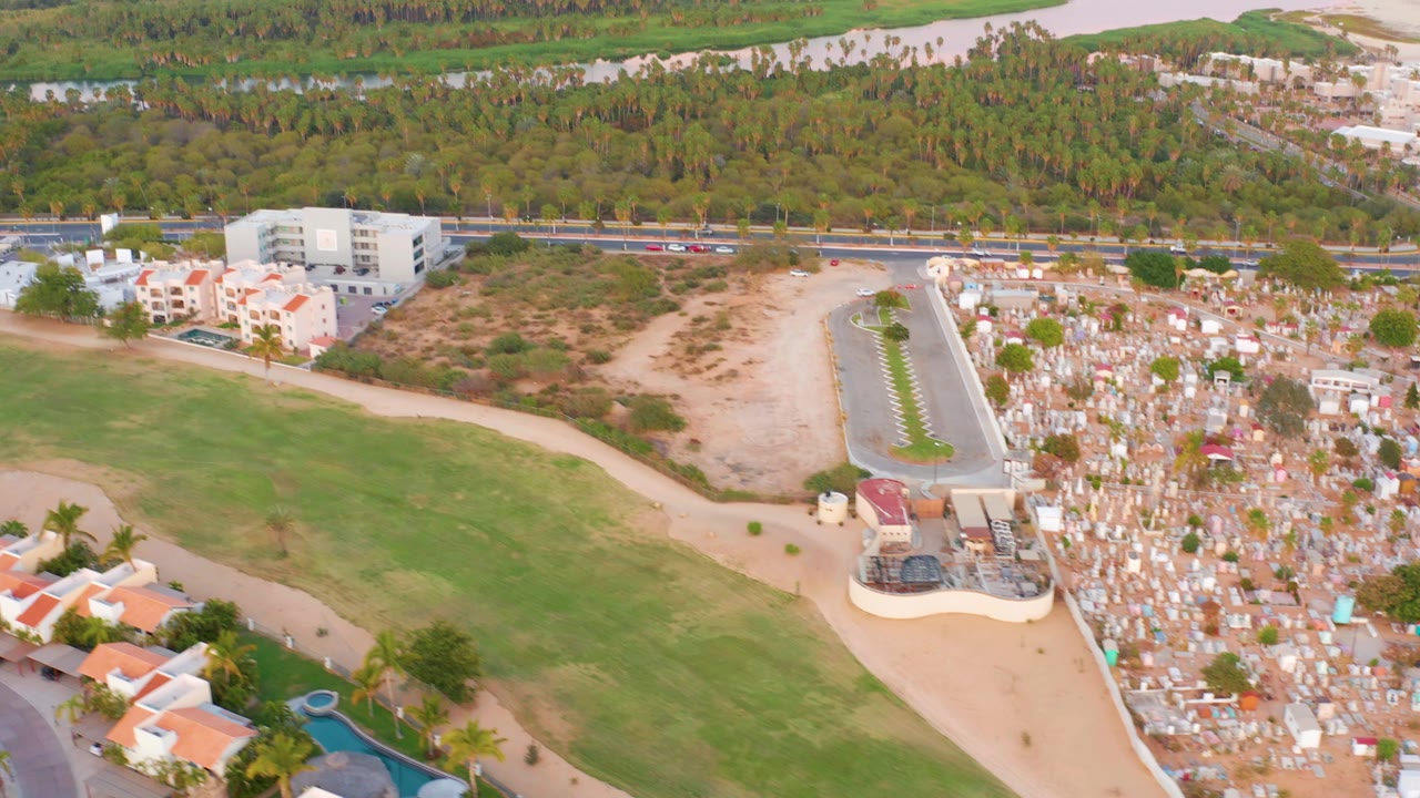 High aerial view of grass field, cemetery plots, residential buildings, road traffic, beach, and water.
