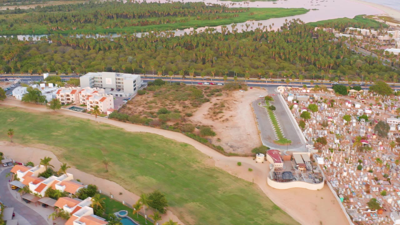 Wide San Jose del Cabo drone frame showing residential blocks and a green field in the foreground, cemetery grounds and moving road traffic in the middle distance, beach and water beyond, and no visible people.